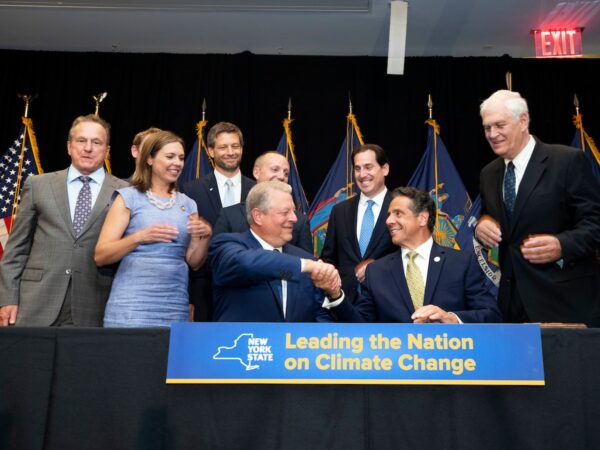 A seated Andrew Cuomo and Al Gore shake hands at a bill-signing ceremony, as a group of people standing behind them excitedly look on