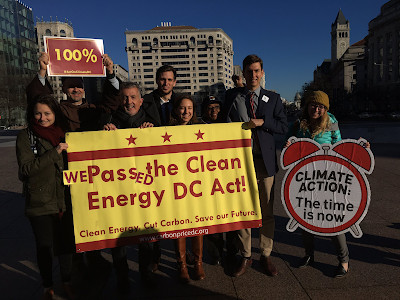 Environmental advocates victoriously hold a big sign that says "We passed the clean energy DC Act!", outdoors in Washington, D.C.