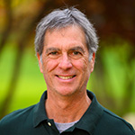 Image of Bill Blancato with short, slightly graying hair, wearing a dark green polo shirt. The background is softly blurred, suggesting a natural setting with greenery.